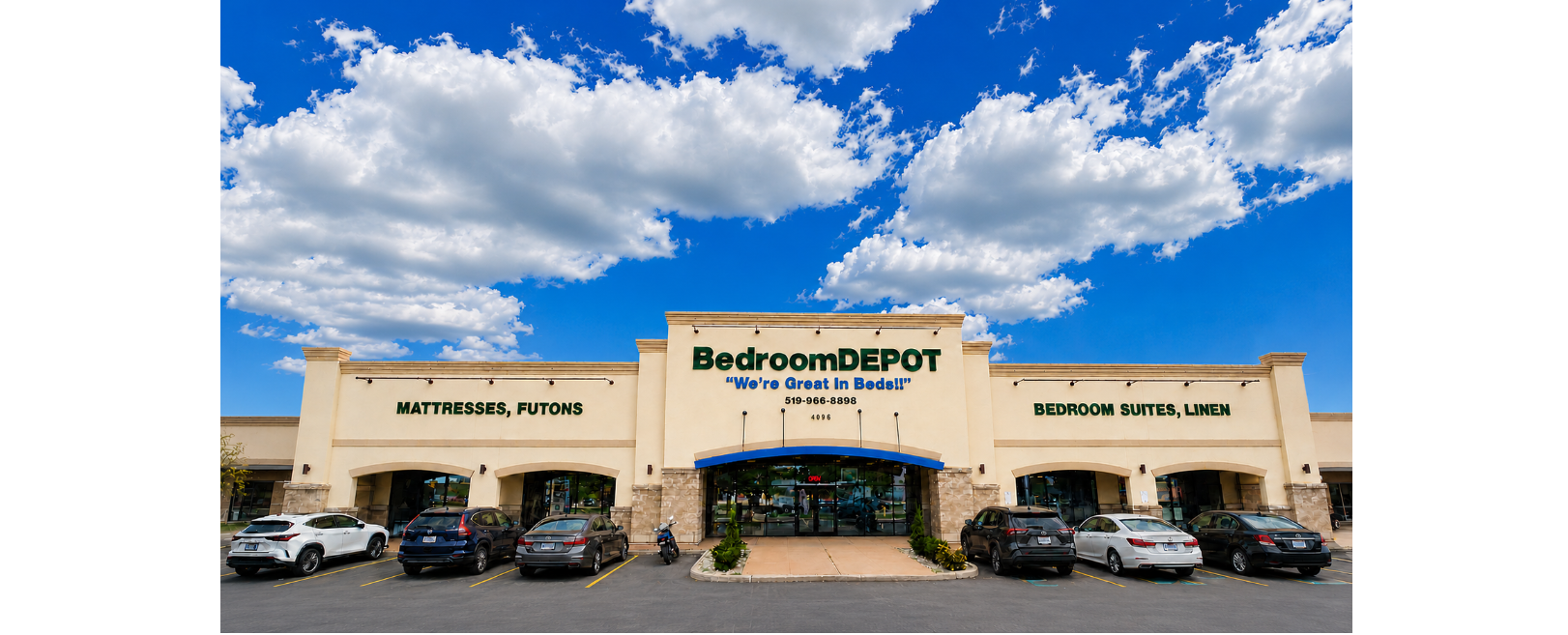 Bedroom DEPOT retail store front with a large beige building, blue awning, parking lot with several cars, and a bright blue sky with white clouds above.